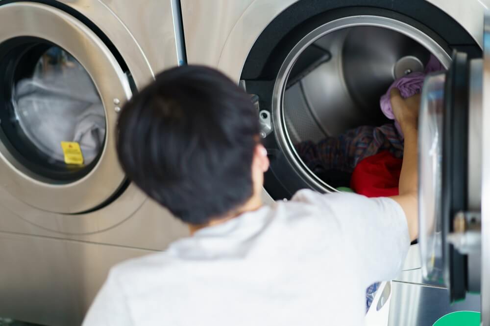 Asian man putting an used or dirty clothes in the self-service automatic laundry washing machine, Asian man using smartphone to access self service KIOS laundry machine.
