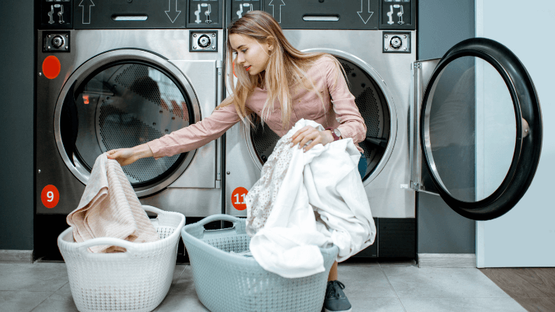 Young and cheerful woman sorting clothes sitting on the dryer machine in the laundry