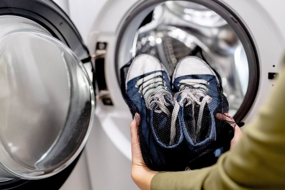 Woman putting blue sneakers in mesh laundry bag into washing machine, close up. Washing dirty sport shoes. Footwear care
