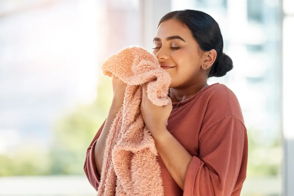 Woman smelling clean laundry, blanket or fabric for fresh and clean smell in house after doing washing, cleaning and housekeeping